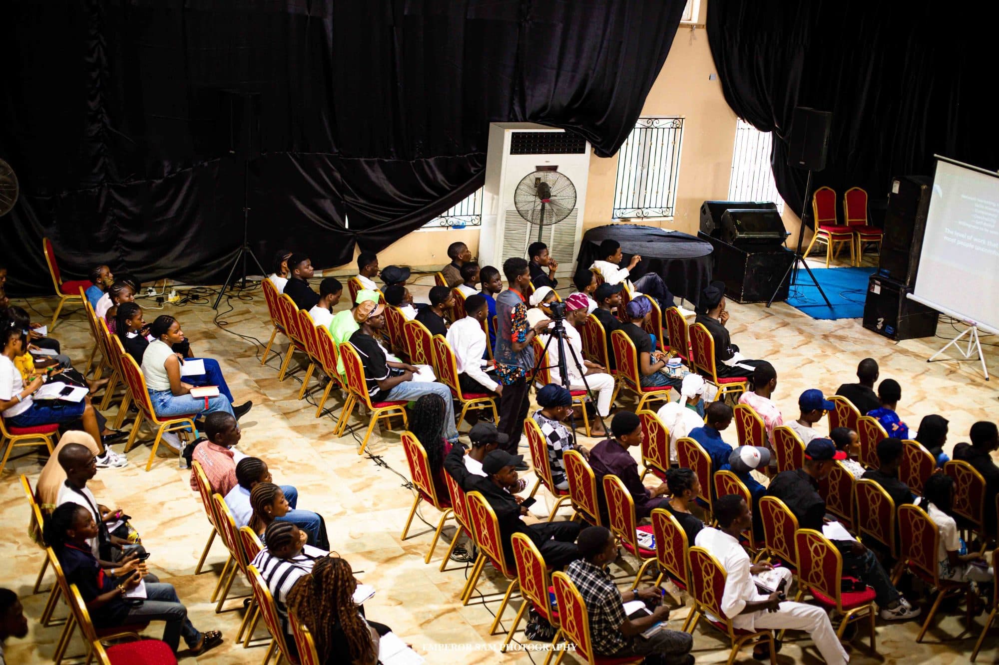 View from above of a full hall of attendees seated in rows facing a presentation screen